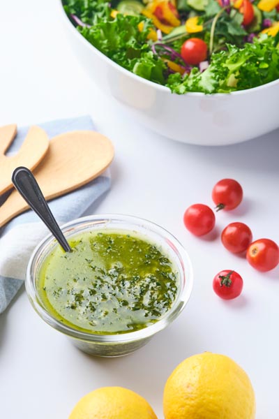 A small bowl with basil pesto vinaigrette inside next to cherry tomatoes, lemons and a bowl of salad.