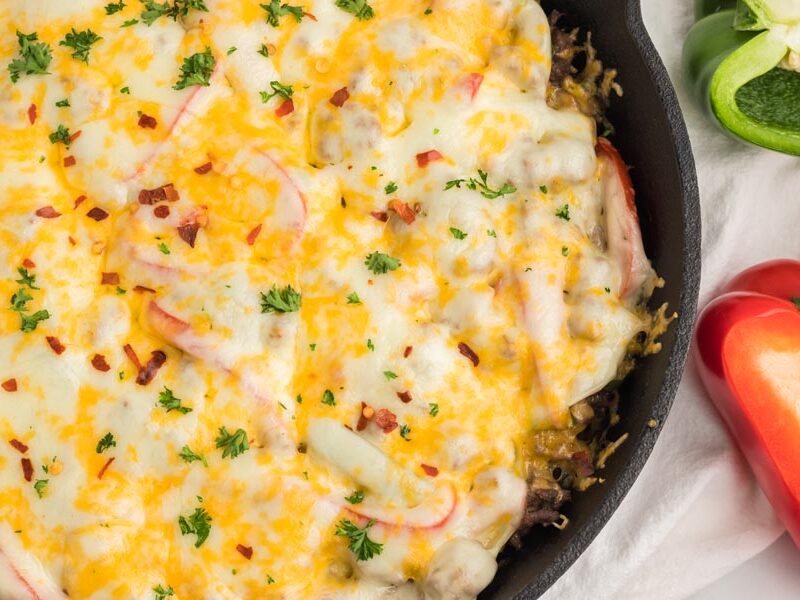 A melted cheese covered skillet casserole dish with sliced red and peppers around.