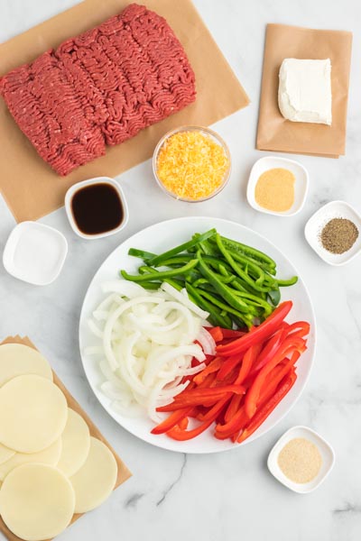 Ingredients for casserole including a plate of raw sliced bell peppers and onion, cream cheese, ground beef, provolone cheese and various spices.