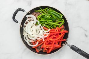 Raw red and green bell pepper and onions sit on top of cooked ground beef in a skillet.