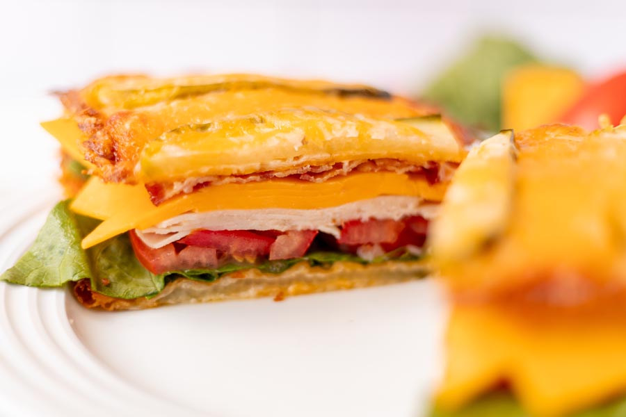 Close-up of a sandwich showing layers of turkey, cheddar, lettuce, and tomato between crispy cheese and pickles.