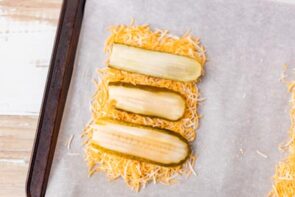 Shredded cheese topped with pickle slices arranged on parchment paper before baking.