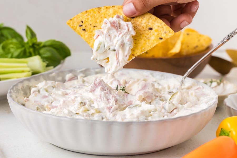 A tortilla chip scooping creamy pickle wrap dip filled with chopped ham and dill pickles.