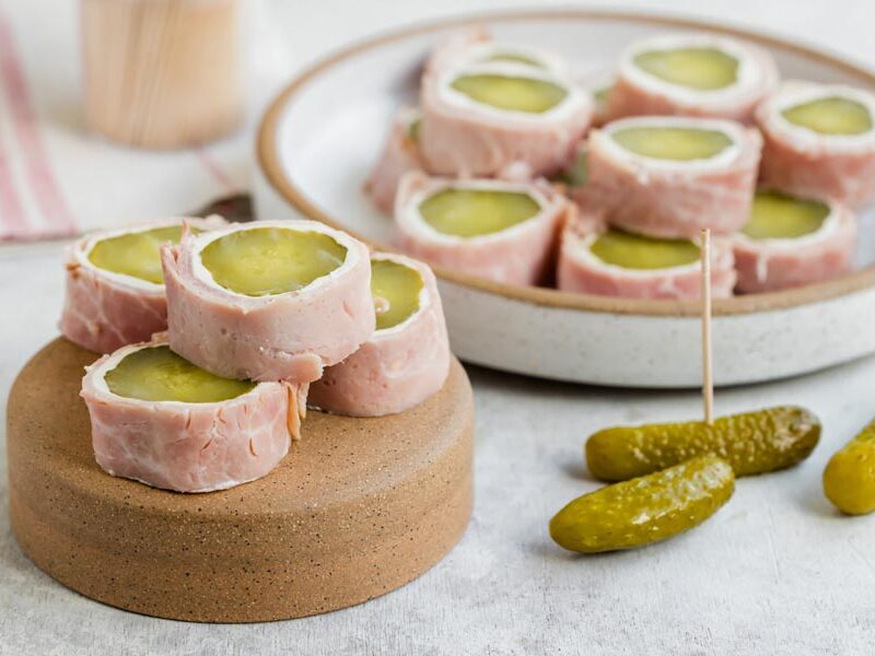 Pickle roll-ups on a small stand and plate, with whole pickles and toothpicks beside them.