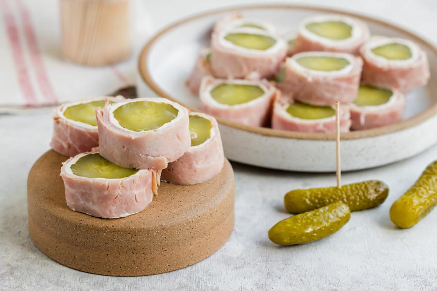 Pickle roll-ups on a small stand and plate, with whole pickles and toothpicks beside them.