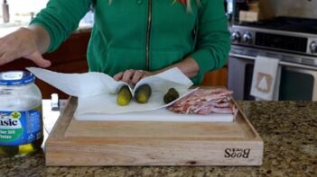 Preparing pickles and deli meat on a cutting board with a jar of pickles and paper towels.