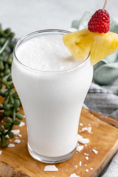 a glass with a white creamy pina colada smoothie inside garnished with pineapple, coconut flakes and a raspberry
