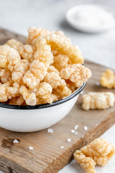 Perfectly fried chicharrones in a bowl on a wooden board with coarse salt nearby.
