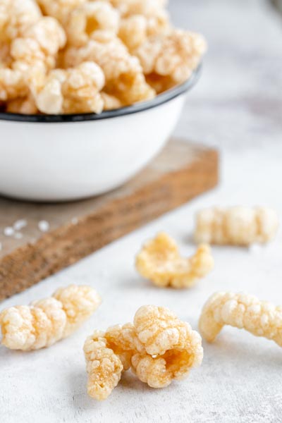 Homemade pork rinds on a table next to a bowl of them on a wood board.