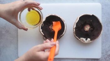 Hands brushing oil with an orange silcone brush onto the gills of two mushroom caps.