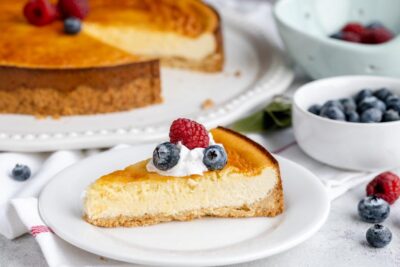 A slice of cheesecake on a plate topped with whipped cream and berries. With a bowl of blueberries next to the plate.