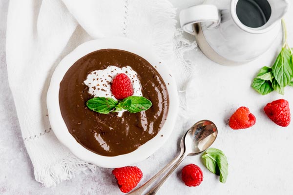 a bowl of chocolate pudding next to mint leaves, raspberries and a creamer container