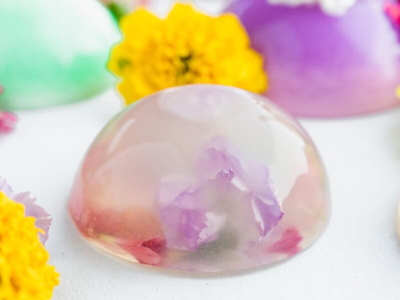 Clear raindrop cake with a purple flower inside, surrounded by colorful flowers.