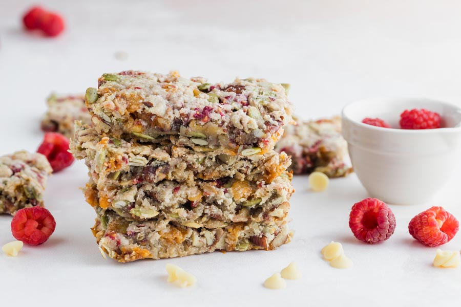 a stack of raspberry granola bars next to white chips and a bowl of fresh raspberries