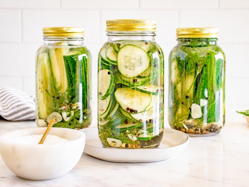 Homemade sliced refrigerator pickles in a mason jar surrounded by jars of dill pickle spears and fresh herbs.