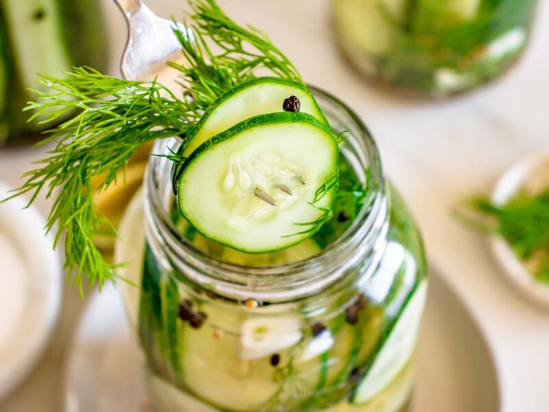Thinly sliced refrigerator dill pickles being lifted from a mason jar with a fork alongside fresh dill and pickling spices.