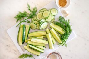 Fresh cucumbers cut into thin rounds and spears on a cutting board with dill, garlic, and mustard seeds for homemade refrigerator pickles.