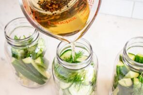 Homemade vinegar pickling brine being poured into a mason jar filled with cucumber slices, garlic, and fresh dill.