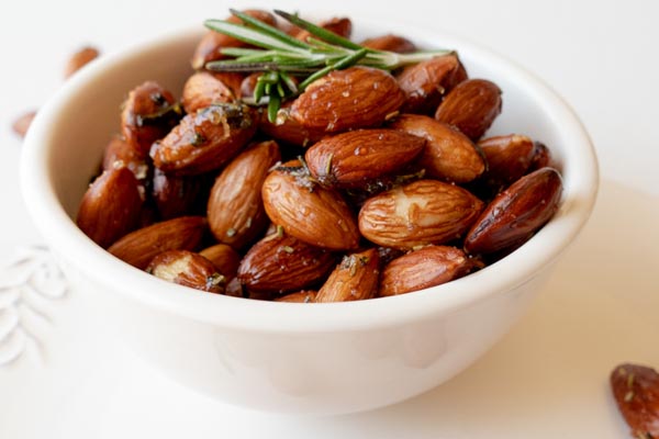 a white bowl filled with roasted almonds and a couple of rosemary leaves on top