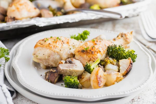 a plate with two chicken legs and baked vegetables on it with a baking tray nearby