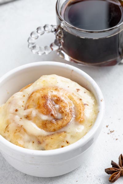 A gooey cinnamon roll in a white ramekin topped with icing sits in front of a cup of black coffee.