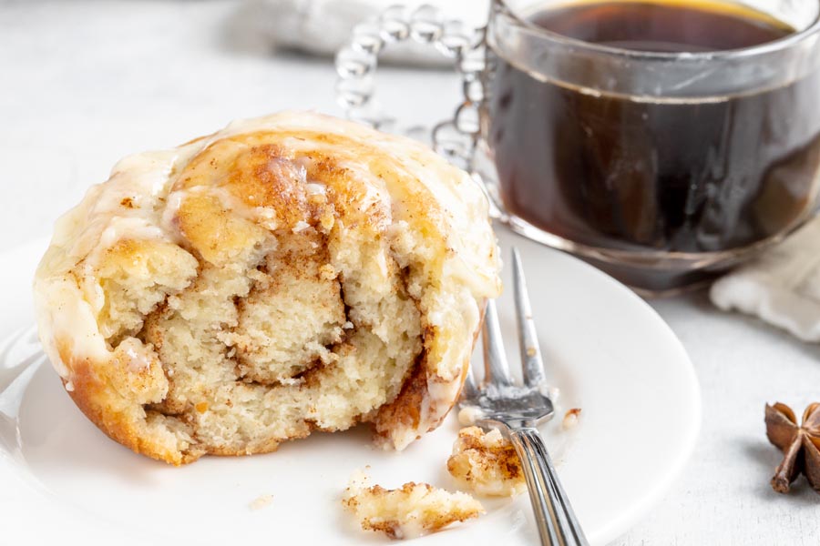 Half of a cinnamon roll on a plate showing the cinnamon swirl layers. A fork sits on the plate with a cup of coffee in a clear glass mug is in the rear.