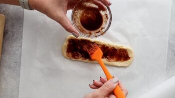 Hands painting a buttery cinnamon mixture on the strip of bread dough with an orange silicone brush.