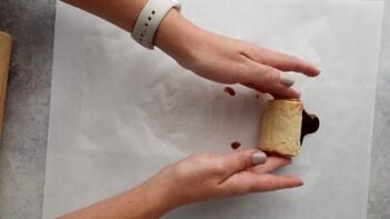 Hands rolling a raw cinnamon roll on a sheet of parchment paper.