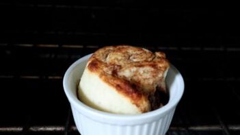 A single cinnamon roll baking in the oven inside a white ramekin.