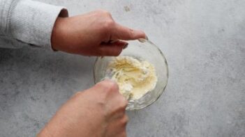 Hands mixing a cream cheese frosting in a clear bowl with a fork.