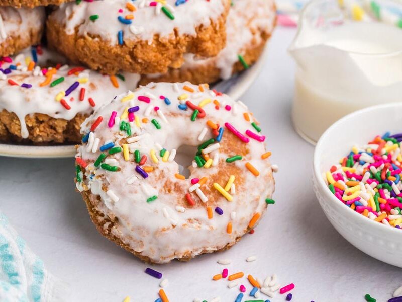 A glazed keto donut standing in front of plate of more donuts and a bowl of colorful sprinkles.