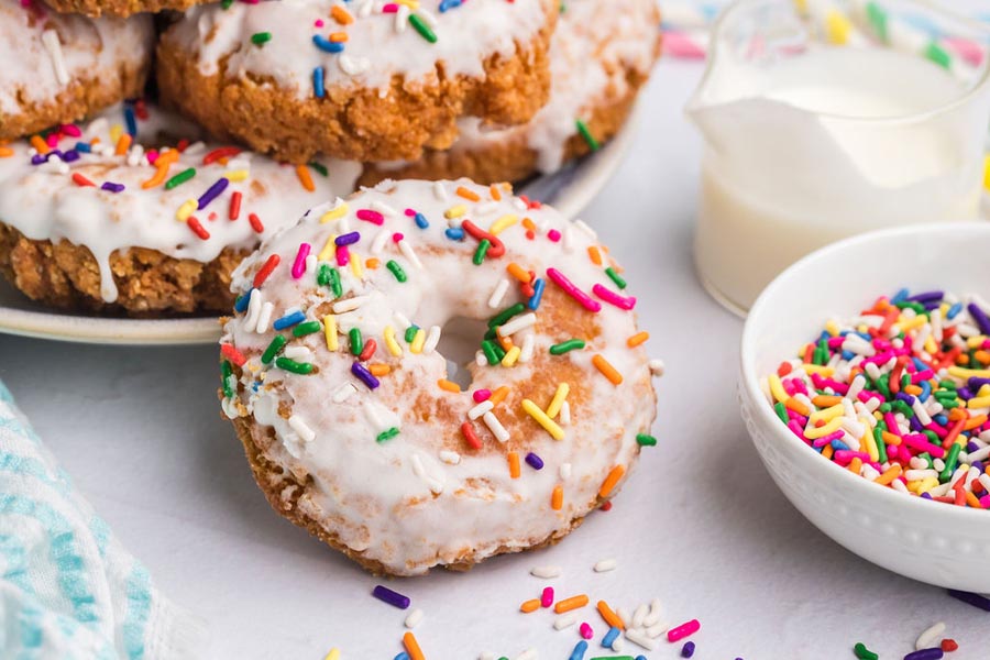 A glazed keto donut standing in front of plate of more donuts and a bowl of colorful sprinkles.