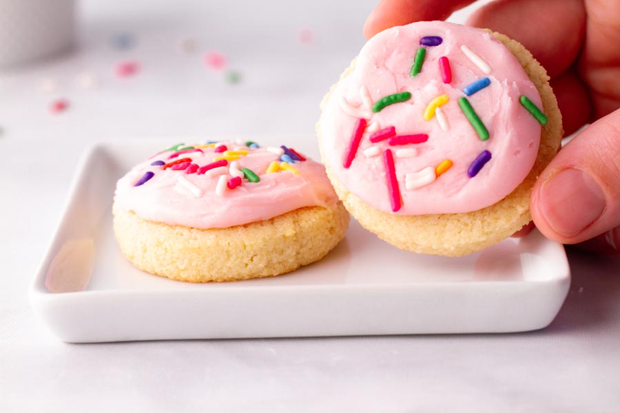 A hand holding a pale pink Lofthouse cookie next to another cookie sitting on a plate.