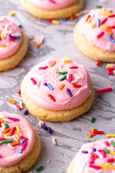 Multi-colored sprinkles cover fresh baked frosted cookies and sprinkles are scattered over the baking tray.