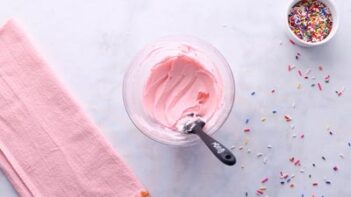 Pale pink buttercream frosting in a glass bowl with sprinkles scattered around.
