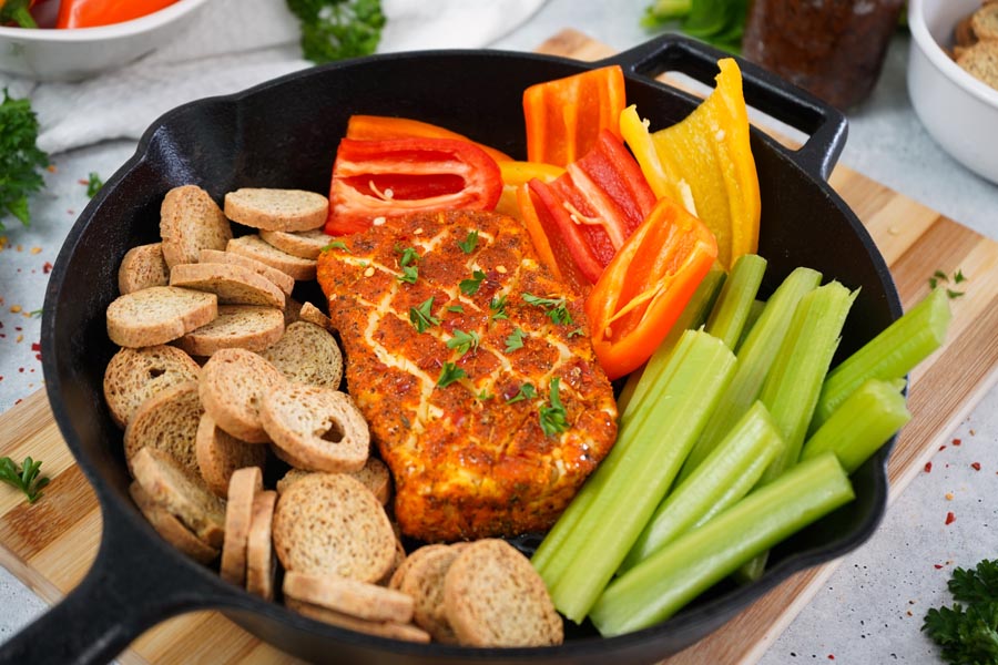 A block of seasoned and smoked cream cheese next to fresh vegetables and melba toasts sitting in a cast iron skillet.