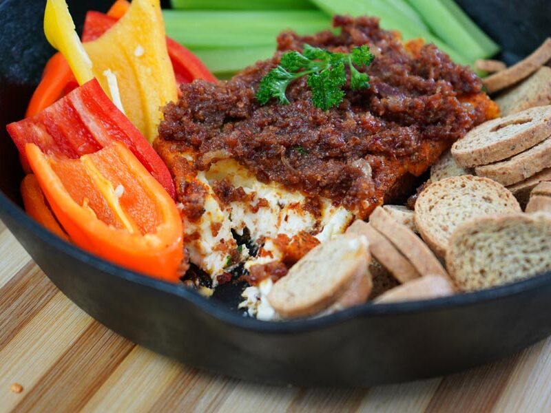 Bacon jam tops smoked cream cheese in a cast iron skillet with crackers and mini sweet peppers to the side.