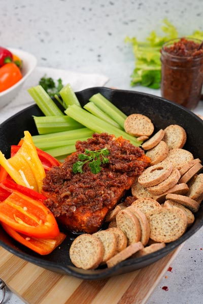 Bacon jam is on top of a block of cream cheese, with melba toast and bell peppers around.