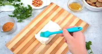 A hand brushing olive oil on a block of cream cheese sitting on a cutting board.