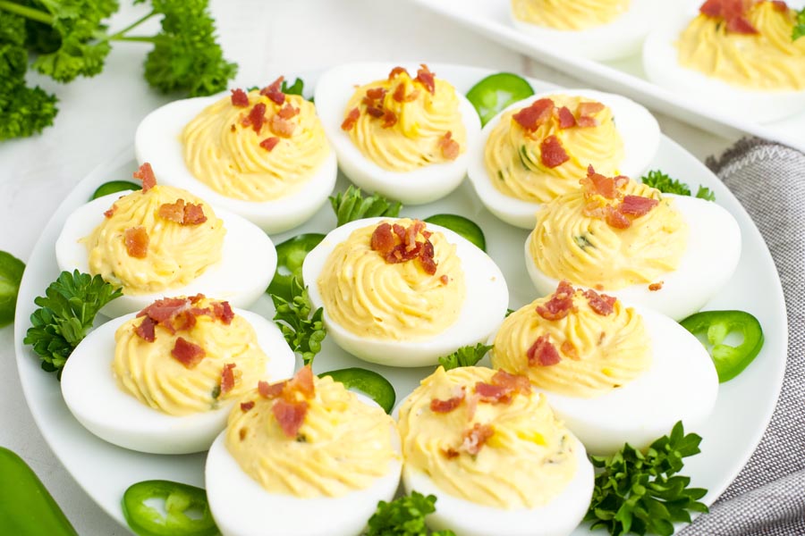 Jalapeno bacon deviled eggs on a plate with sliced jalapenos and parsley surrounding.