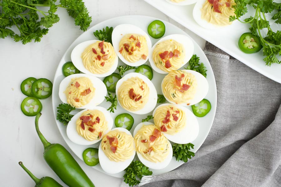 Looking down onto a plate with spicy deviled eggs filled with jalapenos and topped with bacon. Jalapeno slices are nearby.