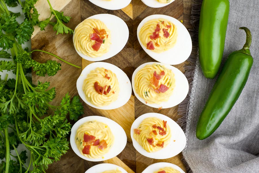 Deviled eggs topped with bacon on a wooden board with parsley and jalapenos nearby.