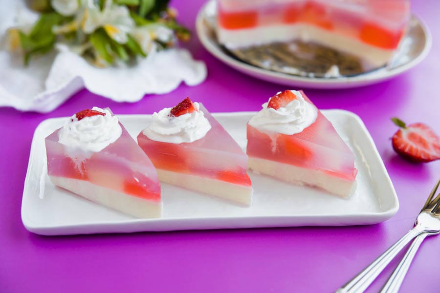 Three triangular slices of stained glass cake on a white tray, each topped with whipped cream and strawberry.