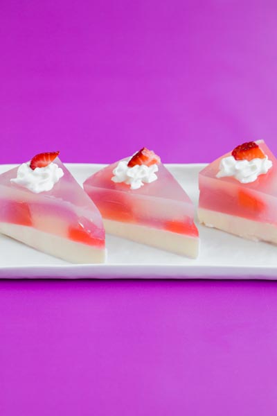 Three triangular slices of stained glass cake with strawberries on a white tray and purple background.