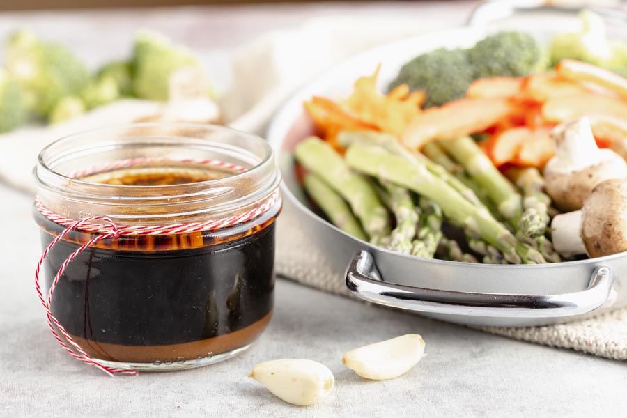 a mason jar with brown stir fry sauce has a red and white string tied around it sits next to a skillet with raw vegetables of asparagus, mushrooms and garlic