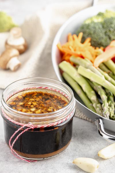 a small jar with homemade stir fry sauce with garlic next to raw asparagus, mushrooms and shredded carrots