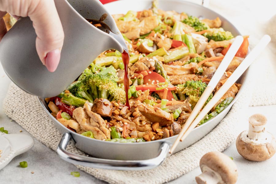 pouring sauce into a skillet with chicken stir fry with chopsticks on the side