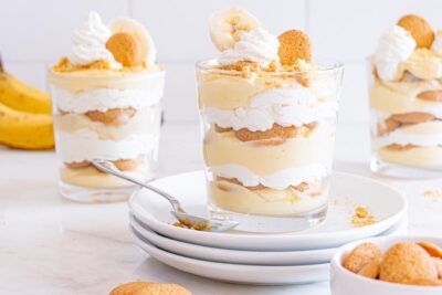 Three banana pudding trifles filled with pudding, whipped cream and vanilla wafers with one on a stack of plates.