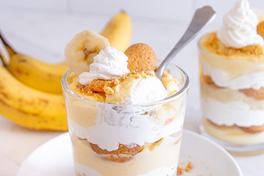 A spoon holding a bite of whipped cream and banana pudding above a trifle dessert.