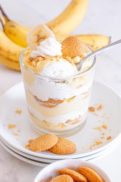 A single southern style banana pudding with a spoon holding a bite over the top of the dish. Bananas are in the background.